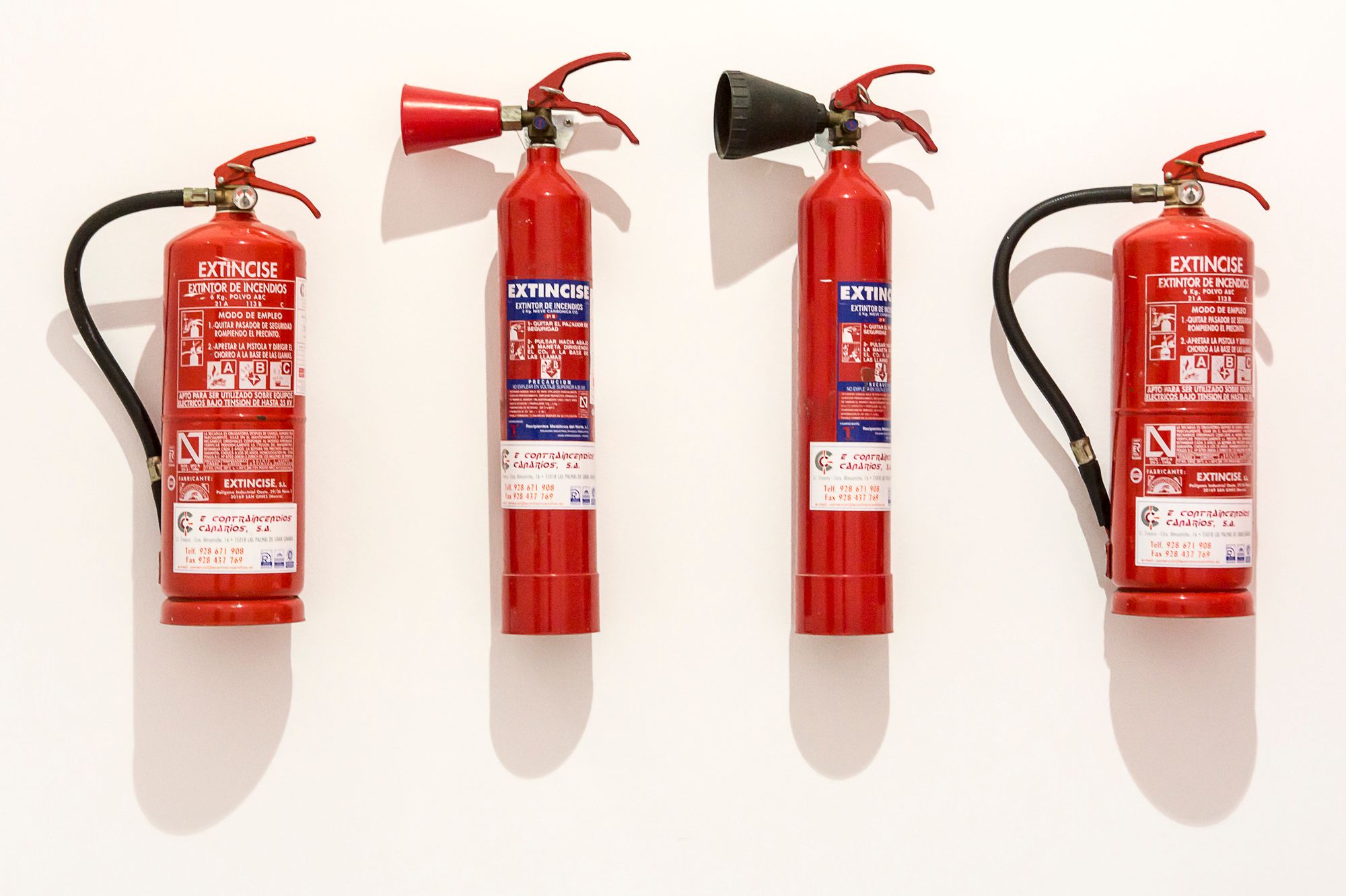 Cuatro extintores rojos de distintos agentes colgados en un muro blanco