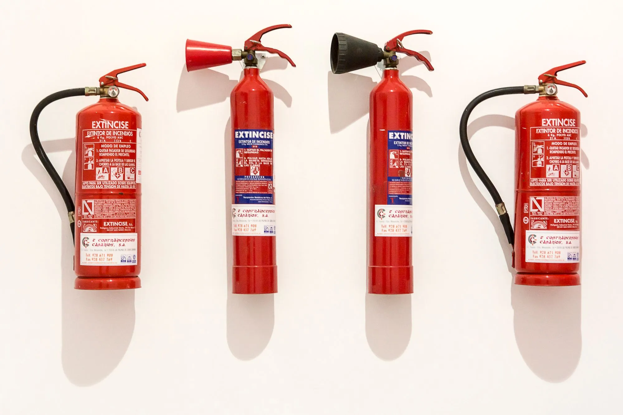 Cuatro extintores rojos de distintos agentes colgados en un muro blanco