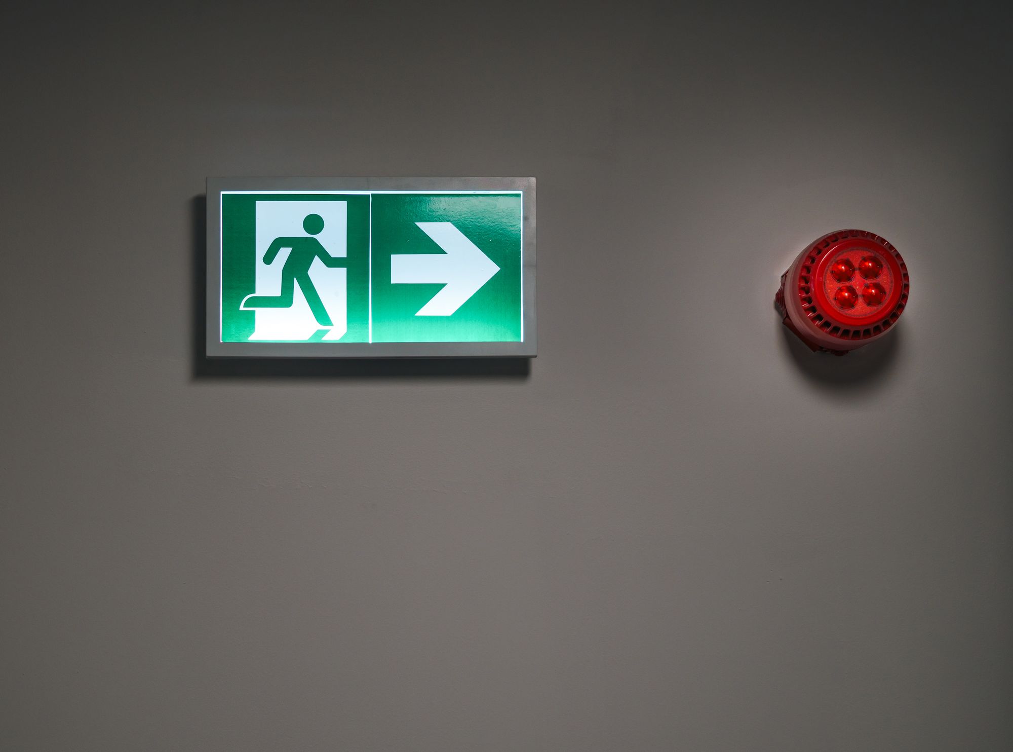 Señal iluminada de salida de emergencia junto a una alarma contra incendio en muro gris