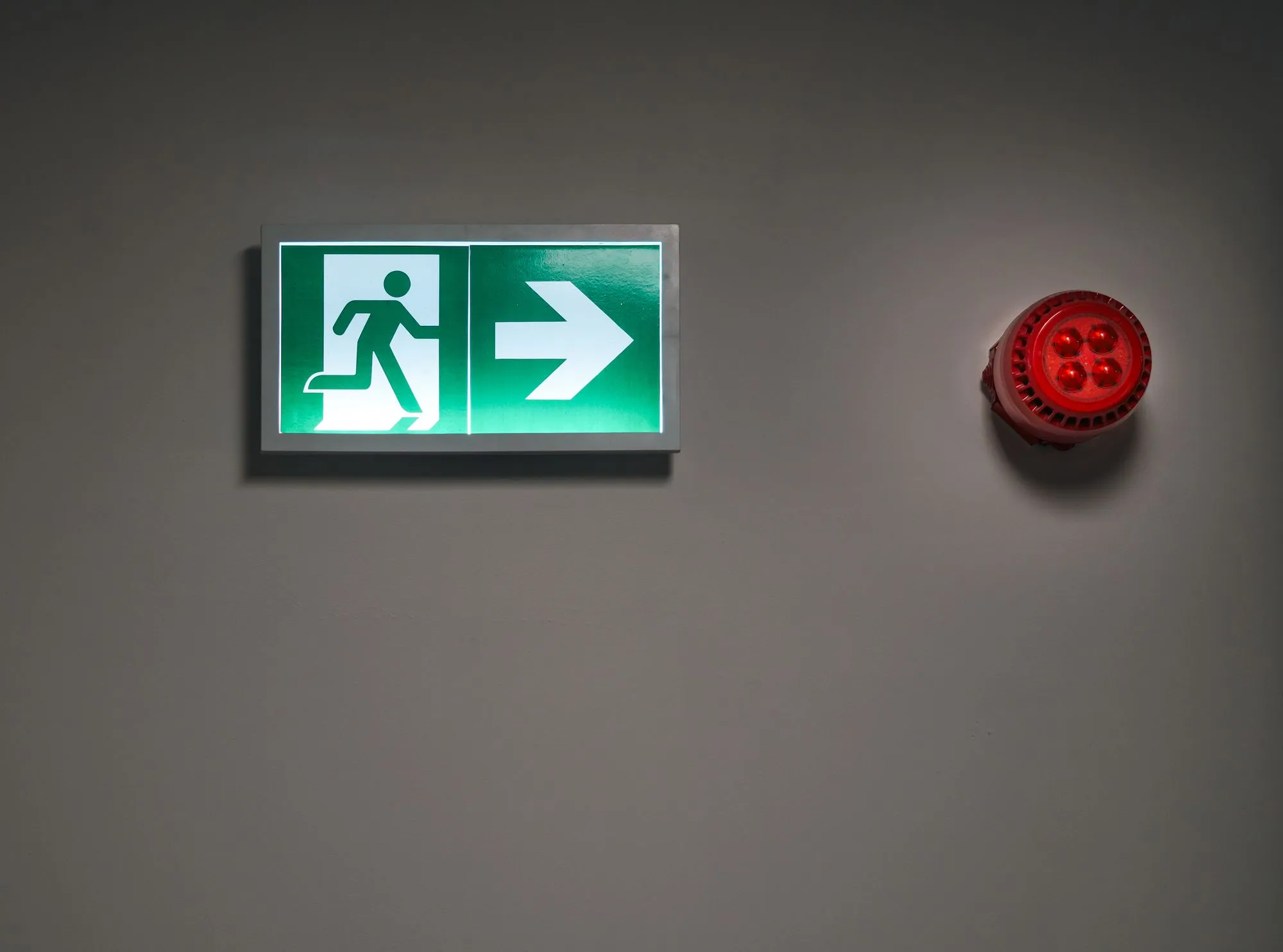 Señal iluminada de salida de emergencia junto a una alarma contra incendio en muro gris