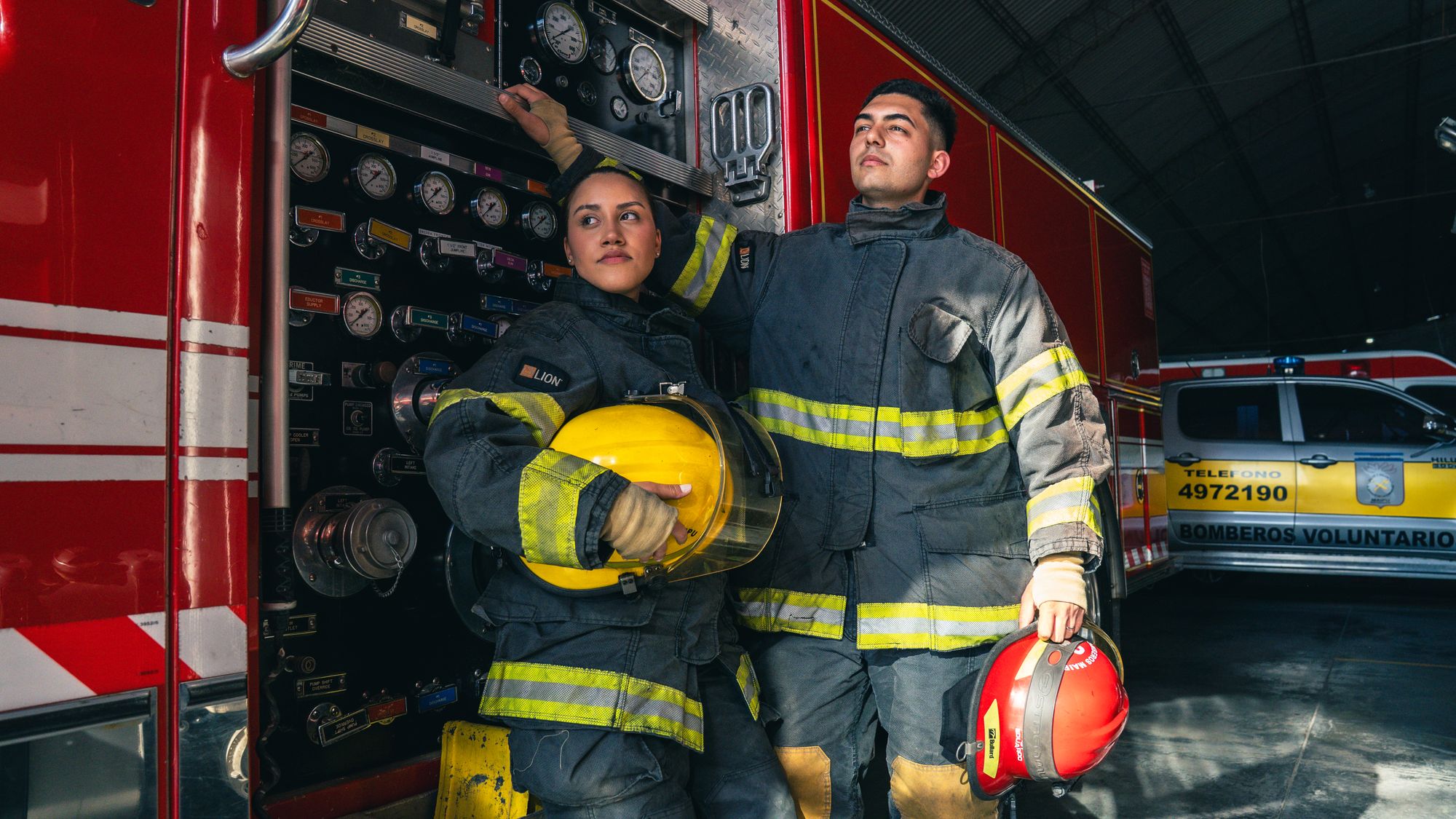 Dos bomberos con traje estructural y casco frente a una unidad de bomberos