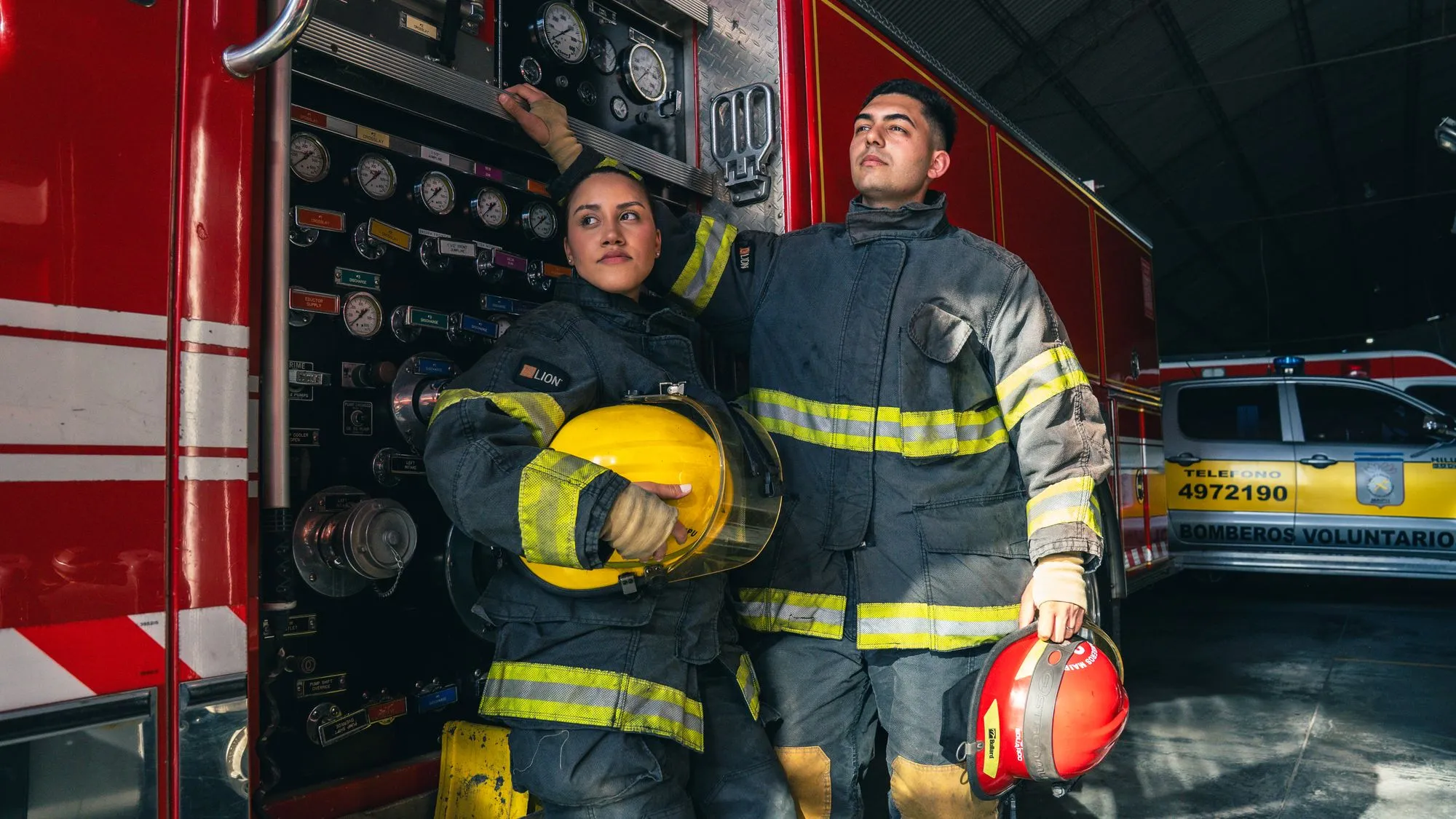 Dos bomberos con traje estructural y casco frente a una unidad de bomberos